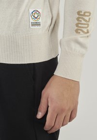 Maglione a maglia beige chiaro con polsini e bordo a coste, caratterizzato da un patch logo multicolore e con la scritta "2026" in oro sulla manica.
