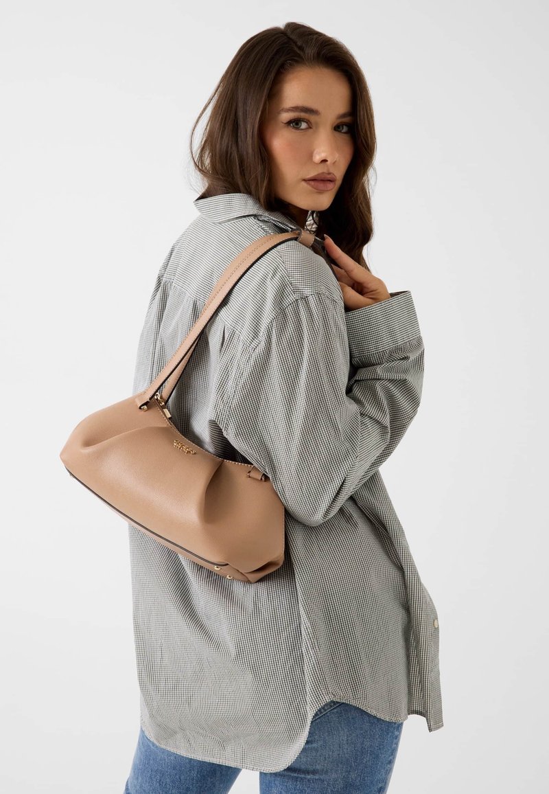 Femme portant une chemise à carreaux grise et un jean bleu, avec un sac à main en cuir beige porté à l'épaule, regardant par-dessus son épaule.