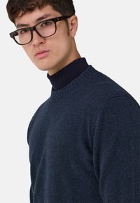 Granatowy sweter z dzianiny z prążkowanym okrągłym dekoltem i dopasowanym krojem, noszony na ciemnym golfie. Widoczna teksturowana tkanina.