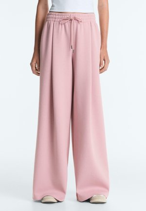 Kobieta ubrana w luźne, szerokie różowe spodnie ze ściągaczem w talii i białe trampki, stojąca na tle jednolitego jasnego tła.