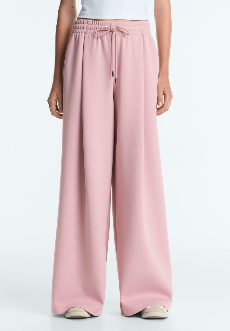 Femme portant un pantalon ample rose à jambes larges avec une taille à cordon et des baskets blanches, se tenant devant un fond clair uni.