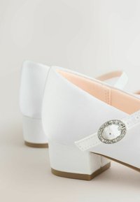Białe satynowe buty na niskim obcasie z zaokrąglonym noskiem, zdobione dekoracyjną sprzączką z kryształkami po boku oraz różową podszywką.