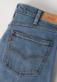 Πίσω τσέπη σε μπλε τζιν Levi’s με πορτοκαλί ραφές, καρφιά, κόκκινη ετικέτα Levi’s και δερμάτινο λογότυπο στη ζώνη.