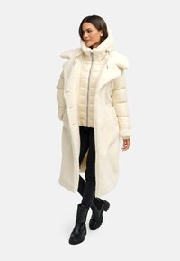 Lang cremet frakke med en blød, tekstureret ydre, lagdelt over en let pufferjakke. Sorte skinny jeans og kraftige sorte støvler fuldender outfittet.