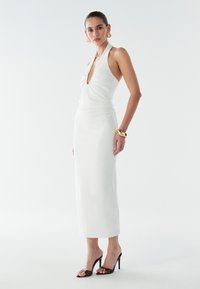 Robe blanche à col halter avec un décolleté en V profond et des détails froncés. Tissu lisse, silhouette ajustée et ourlet fendu. Portée avec des sandales à talons noires.