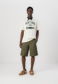 Creme t-shirt med grøn print, olivenfarvede cargo shorts og tan sko. En sort bandana bæres om halsen. Casual tøj med en afslappet pasform.