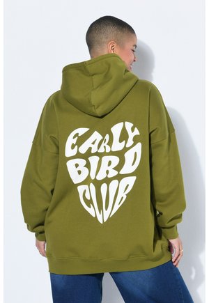 Osoba o krótkich włosach ubrana w oliwkowozieloną bluzę z kapturem z napisem "EARLY BIRD CLUB" w białym kształcie serca na plecach.