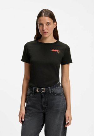 Kvinde iført en sort t-shirt med en lille spyd af røde kirsebær på brystet, kombineret med mørke høj-taljede jeans og et sort bælte.