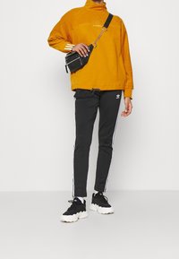 Orange fleece-tröja med hög krage, svarta träningsbyxor med vita ränder, och svarta sneakers med vita sulor. Liten svart crossbody-väska.