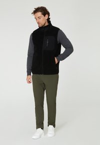 Svart fleeceväst över en grå långärmad skjorta, med olivgröna byxor och vita sneakers. Har en bröstficka och dragkedjeslutning.