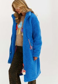 Modrá nadýchaná bunda s textúrovaným povrchom, vybavená zipsom, dvoma bočnými vreckami a dekoratívnym príveskom v tvare huby. Nosená cez vzorovaný sveter.