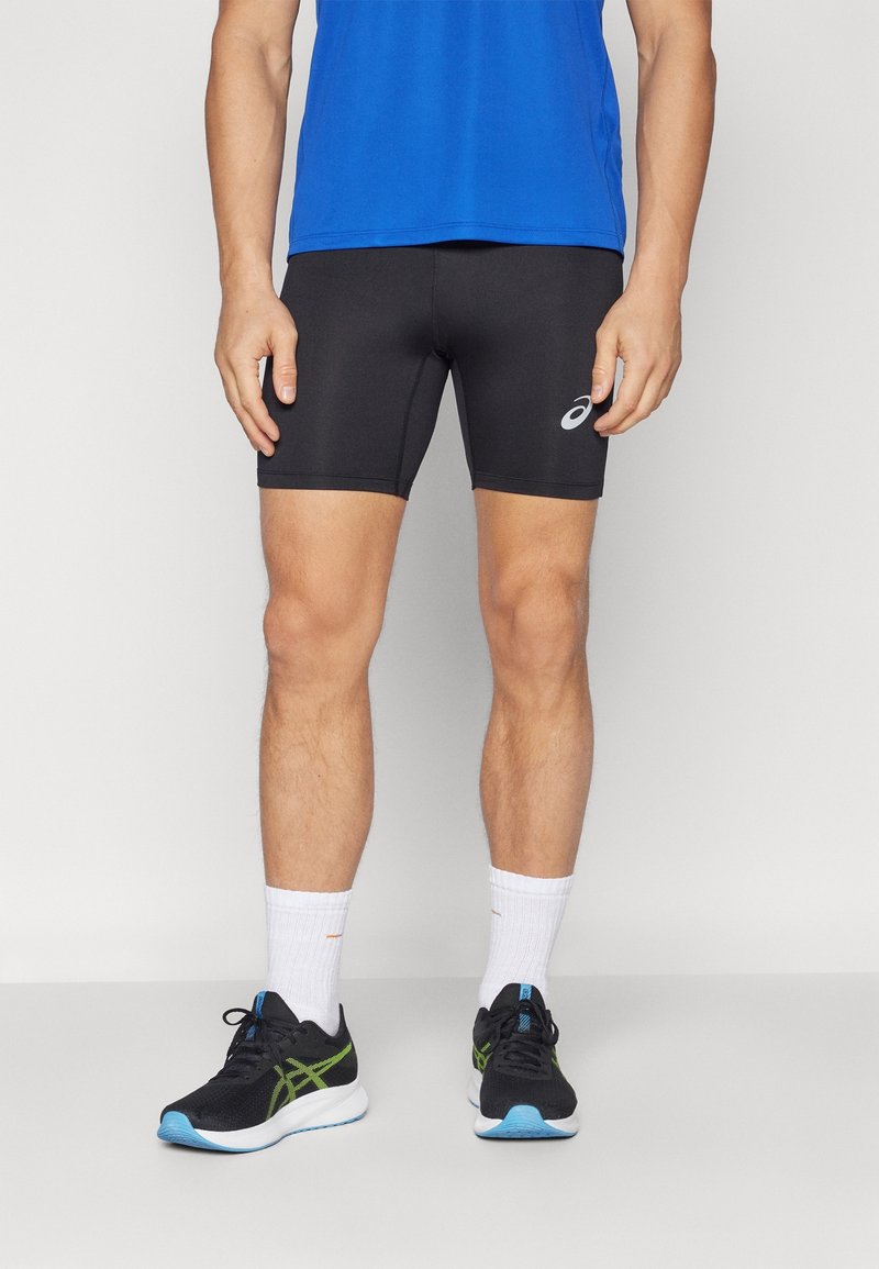 Homme portant un short de sport noir, un t-shirt bleu, des chaussettes blanches et des chaussures de course noires avec des accents verts et bleus, debout sur un sol gris.