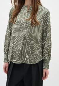 Blusa verde a fantasia in tessuto leggero con chiusura a bottoni, maniche texturizzate e linee ondulate bianche su sfondo oliva.