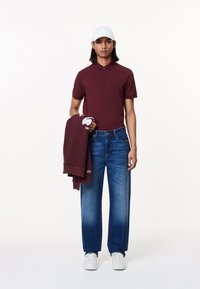 Mörkröd polo skjorta, blå denim jeans med en avslappnad passform, och vita sneakers. Håller en mörkröd sweatshirt med logotypdetaljer och bär en vit keps.