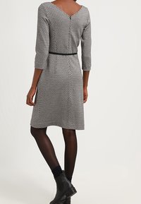 Robe grise à motifs avec un dos en V, manches trois-quarts, taille cintrée et ceinture. Portée avec des collants noirs et des bottines.