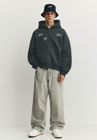 Sötétszürke cipzáras kapucnis pulóver grafikus hímzéssel, világosszürke bőszárú farmerrel és fehér tornacipővel párosítva. A szettet egy fekete kötött kalap egészíti ki.