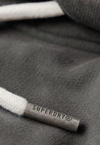 Primo piano di un tessuto grigio con un cordino bianco dotato di un terminatore in metallo testurizzato etichettato "SUPERDRY" ben in evidenza.