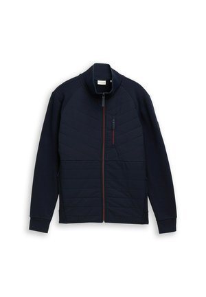 Marine blauwe zipjack met een gequilt voorpaneel, een gladde achterkant, hoge kraag en rode ritsaccenten. Bevat zijzakken en gestructureerde stof.
