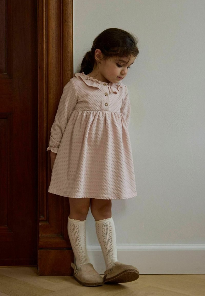 Jeune fille en robe rose texturée et chaussettes hautes en tricot crème, debout près d'une porte en bois, regardant vers le sol.