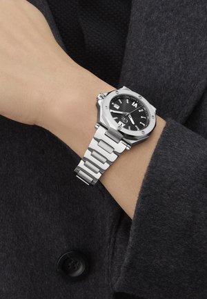 Srebrna uro iz nerjavečega jekla z črnim številčnico, osemkotno ohišje in povezano zapestnico. Prikaz vključuje oznake ur in okno za datum.