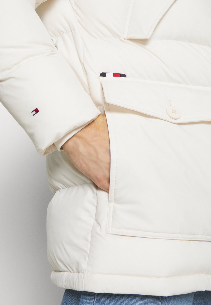 Chaqueta acolchada crema con textura suave, gran bolsillo frontal y detalle de logo bordado en la manga. Presenta un cierre de botones.