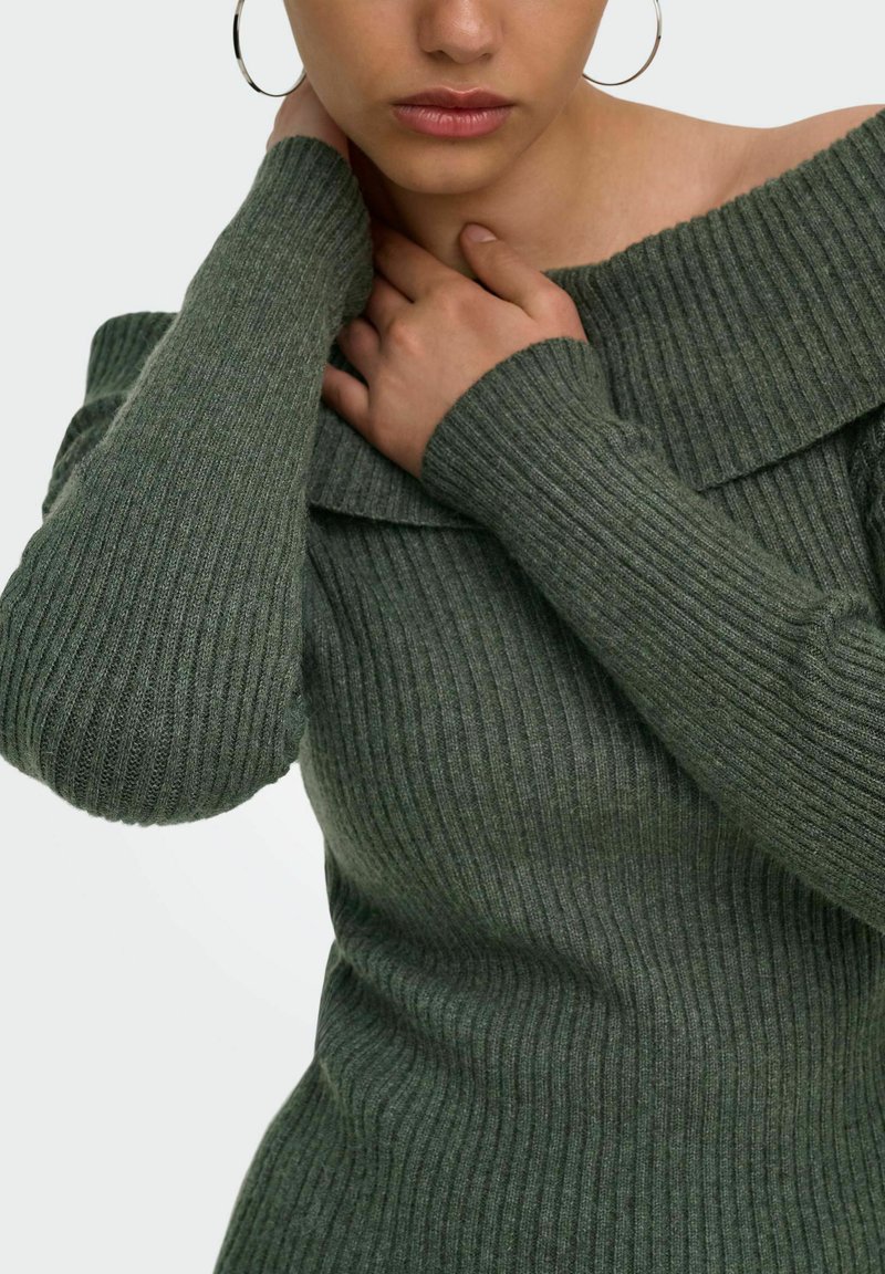 Femme portant un pull vert côtelé à épaules dénudées, tenant son cou d'une main et touchant le col de l'autre.