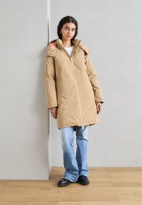 Beige polstret parka med høy krage, gullglidelås og sidelommer. Brukt over lyseblå vide jeans og svarte flate sko.