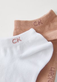 Dos pares de calcetines tubular: uno blanco y uno marrón claro. Cada uno presenta una textura acanalada y un pequeño logotipo "CK" en un color a juego en el puño.