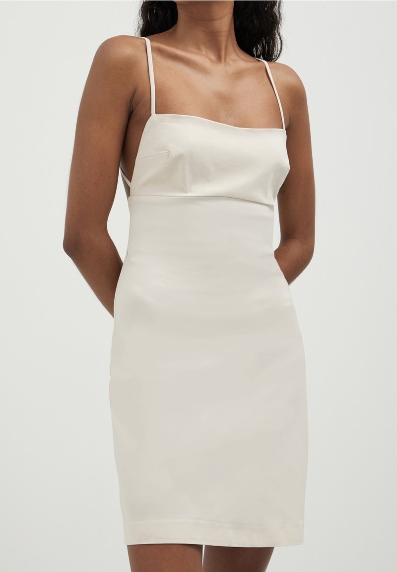 Femme portant une robe blanche moulante sans manches avec de fines bretelles, montrée du cou jusqu'à mi-cuisse sur un fond uni.