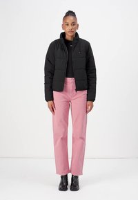 Svart pufferjacka med hög krage, parat med ljusrosa rakbenjeans och svarta ankelskor. Modellen står mot en vit bakgrund.