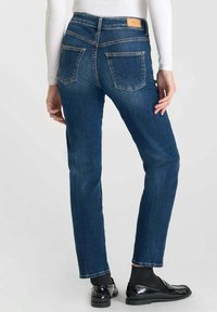 Jean en denim bleu foncé avec une coupe droite, taille mi-haute et deux poches arrière. Présente des coutures discrètes et une étiquette en cuir beige.