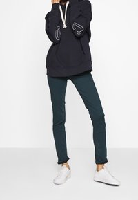 Marineblauer Hoodie mit weißen Akzenten und Kordelzug, kombiniert mit dunkelgrüner Hose und weißen Sneakern. Angenehme Textur und entspannte Passform.