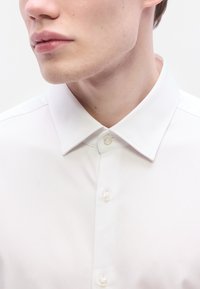 Λευκό πουκάμισο με κουμπιά και κλασικό γιακά. Διαθέτει λεία υφή και διακριτική γυαλάδα. Δύο κουμπι ά εμφανίζονται στο λαιμό.