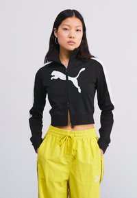 Black cropped zip-up jacket with white Puma logo, featuring high collar, contrasting white side stripes, worn with yellow drawstring pants.