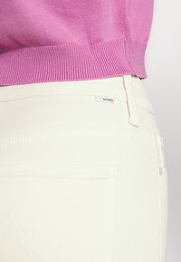 Suéter de punto rosa claro combinado con jeans crema. Los jeans cuentan con una etiqueta de logo sutil y un diseño distintivo de costura en los bolsillos.