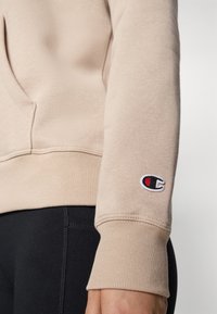 Sweatshirt beige avec une finition texturée, doté d'une poche kangourou et de poignets côtelés. Patch logo notable sur la manche gauche.