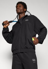 Veste de piste légère noire avec taille et poignets élastiques, arborant un logo sur la poitrine, assortie à un pantalon. Raquette de tennis tenue à la main.