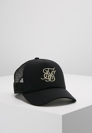 Casquette de camionneur noire en mesh avec visière incurvée et initiales stylisées beige brodées sur le devant, posée sur une surface blanche contre un fond neutre.