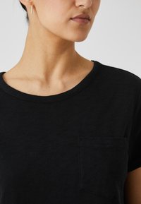 Svart t-skjorte med kort erme som har rund hals og en liten lomme på venstre side, laget av mykt, teksturert stoff.