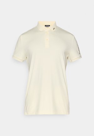 Polo en crème clair, à manches courtes, avec une patte de boutonnage à trois boutons, logo sur le col et des coutures décoratives sur les manches. Tissu lisse et léger.