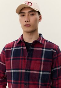 Ung mand iført en beige kasket og en rød og navyblå ternet skjorte over en sort t-shirt mod en lys baggrund.