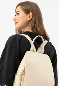 Béžový kožený batoh s strukturovaným designem, zaobleným vrcholem a texturovaným povrchem. Má nastavitelná ramenní popruhy a malou přední kapsu se zipem.