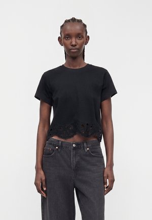 Kvinde med fletninger iført en sort kortærmet cropped top med blonderkant og mørkegrå jeans med høj talje mod en ensfarvet baggrund.