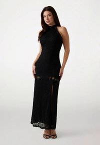 Sort blonde aftenkjole med en tætsiddende silhuet, høj hals og sideslids. Har indviklede blomstermønstre og gennemsigtige detaljer.