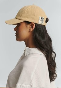 Khaki pet met een gebogen rand, gemaakt van katoen. Bevat een zijlabel met tekst en een verstelbare band aan de achterkant.