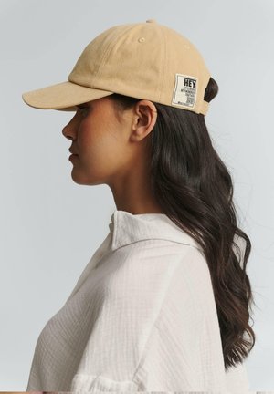 Khaki baseball cap with a curved brim, made of cotton. Features a side label with text and a back adjustable strap.