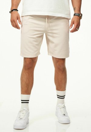 Rippende beigefarbene Shorts mit Kordelzugbund, umgeschlagenen Säumen und lockerer Passform. Getragen mit weißen Turnschuhen und gestreiften Crew-Socken.