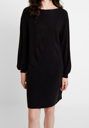 Femme portant une robe en maille noire ample à manches longues avec poignets et ourlet côtelés, debout devant un fond clair uni.