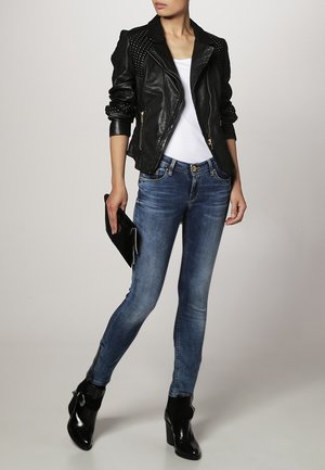 Giacca da motociclista in pelle nera con spalle borchiate, T-shirt bianca, jeans skinny blu e stivaletti neri con tacco alto. Tiene una clutch nera.