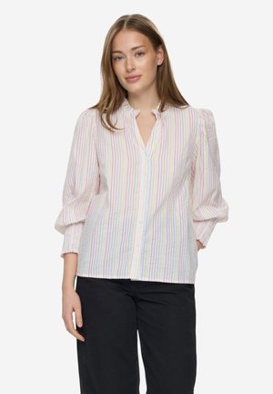 Młoda kobieta ubrana w pastelową, pasiastą bluzkę na guziki z bufkowatymi rękawami oraz czarne spodnie, stojąca na tle jednolitego białego tła.